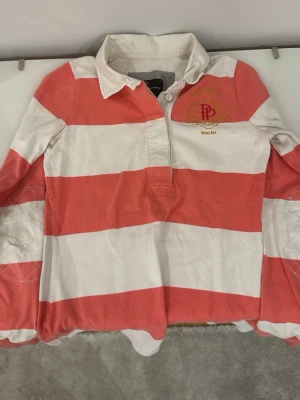 Randig rugbytröja från Polo Ralph Lauren - Säljer en långärmad rugbytröja från Polo Ralph Lauren med breda ränder i rött och vitt. Skjortan har klassisk krage, knappar vid halsen och broderad logga på bröstet. Perfekt för en sportig och avslappnad stil.
