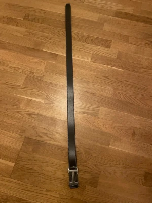 Svart skärp med silverfärgat  h spänne - Snyggt svart bälte i skinn med vit kontrastsöm och ett stort, silverfärgat h -format spänne. Perfekt accessoar för att lyfta din outfit med en clean och stilren look.