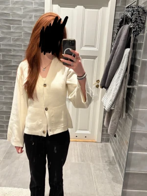 Vit stickad kofta med knappar - Snygg vit stickad kofta i bomull med v-ringning och tre stora knappar framtill. Koftan har långa, lite vidare ärmar och ribbade detaljer vid midjan och ärmsluten. Perfekt att slänga över en topp för en clean och fräsch look. Endast testad