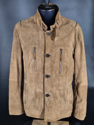 Brun mocka skinnjacka med ståkrage - Snygg brun skinnjacka i mocka med ståkrage och knäppning framtill. Två vertikala bröstfickor med dragkedja och två sidofickor. Jackan har en modern passform och stilrena detaljer som ger en cool look.