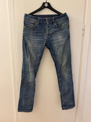 Blå raka jeans från Nudie Jeans W32 L34 - Snygga blå jeans från Nudie Jeans med klassisk femficksdesign och raka ben. Jeansen har slitningar och kontrastsömmar, samt läderpatch bak i midjan. Tillverkade i denim med en autentisk tvättad look.