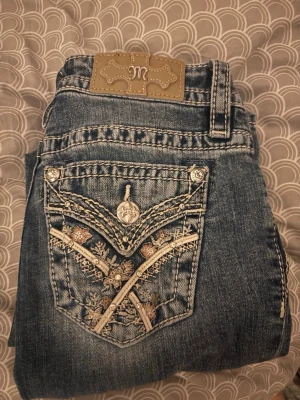 Miss Me jeans med broderade fickor - Säljer ett par blå Miss Me jeans med snygga broderade bakfickor i beige och silverdetaljer. Jeansen har normal passform och klassisk femficksdesign. Perfekta för dig som gillar detaljer och unik stil!