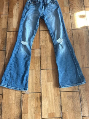 Blå bootcut jeans med slitningar från Gina Tricot  - Säljer ett par ljusblå bootcut jeans med slitna detaljer på knäna och broderade bakfickor med röda detaljer. Endast använda en gång. Säljer pågrund av att dem inte kommer till användning. Storlek 164 från Gina Young. 