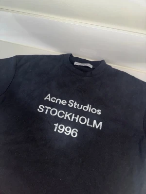 Svart Acne Studios t-shirt Stockholm - Skön svart t-shirt från Acne Studios med vit text 'STOCKHOLM 1996' på. Klassisk rund hals och rak passform. Perfekt för dig som gillar stilrena och trendiga plagg med streetkänsla.