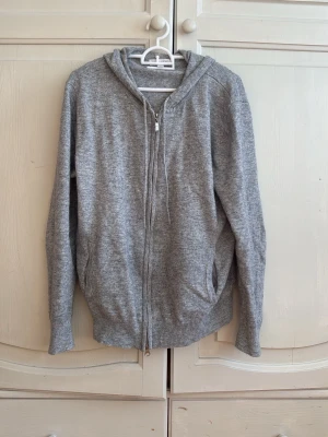 Grå zip hoodie från Ballucio - Mysig grå hoodie från Ballucio med dragked blixlås framtill och två fick do på sidorna. Tillverkad i mjukt stickat material och har ribbade muddar vid ärmslut och nederkant. Perfekt för en avslappnad stil och enkel att match pan med andra almost. Kashmir och ullblanding