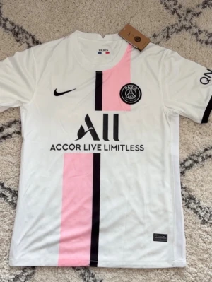 PSG vit matchtröja Nike - Säljer en vit Paris Saint-Germain matchtröja från Nike med rosa och svart vertikal rand på framsidan. Tröjan har PSG-logga på bröstet, sponsortryck och korta ärmar. Materialet är lätt och andas, perfekt för fotboll eller träning.