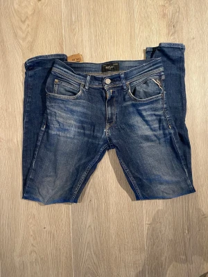 Blå Replay jeans med slitningar - Säljer ett par blå jeans från Replay med snygga slitningar och tvättade detaljer. Klassisk femficksmodell med silverfärgad knapp och dragkedja. Jeansen har raka ben och kontrastsömmar på bakfickorna. Perfekta för en avslappnad och trendig look.