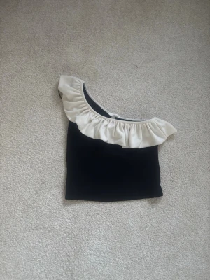 Svart oneshoulder topp med volang - Svart oneshoulder topp med en vit volang från h&m i storlek S. Knappt använd därför väldigt bra skick.