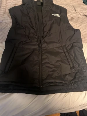 Svart dunväst från The North Face - Snygg svart dunväst från The North Face med dragkedja framtill och två fickor med dragkedja. Västen har en stilren design och är perfekt för lager-på-lager. Tillverkad i slitstarkt material och har logga på bröstet. Perfekt nu inför våren.