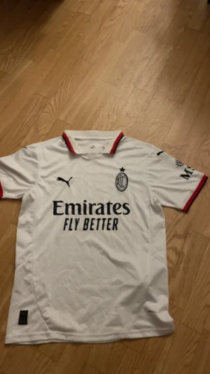 AC Milan vit fotbollströja Puma - AC Milan fotbollströja i vit polyester med svarta och röda detaljer på krage och ärmslut. Puma-logga på bröst och ärm, samt klubbmärke och sponsortryck. Klassisk passform med korta ärmar och snygg krage. Perfekt för dig som älskar italiensk fotboll.