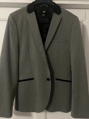 Grå kavaj med svarta detaljer H&M - Stilren grå kavaj från H&M med svarta sammetsdetaljer på kragen och fickorna. Kavajen har en knapp framtill, smal passform och randigt innerfoder med etiketten 'everyday'. Perfekt för en dressad look.