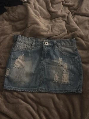 Blå jeanskjol med slitningar - Säljer en blå jeanskjol i kort modell med slitna detaljer framtill och bakfickor. Kjolen har klassisk femficksdesign, bälteshällor och knappstängning i midjan. Perfekt för en avslappnad och trendig look.