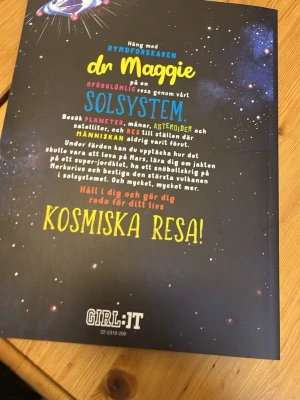 Dr Maggies stora resa genom solsystemet - En färgstark och inspirerande bok om solsystemet skriven av Dr Maggie Aderin-Pocock. Boken har ett svart omslag med illustrationer av planeter, rymdfarkoster och astronaut. Perfekt för dig som vill utforska rymden och lära dig om planeter, månar och asteroider.