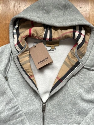 Grå hoodie från Burberry med dragkedja passar både L och M - Snygg grå hoodie från Burberry med dragkedja och klassiskt rutigt foder i huvan. Broderad Burberry-logga på bröstet och mjukt material som känns lyxigt. Perfekt för dig som gillar stilrena detaljer och exklusiva märken.