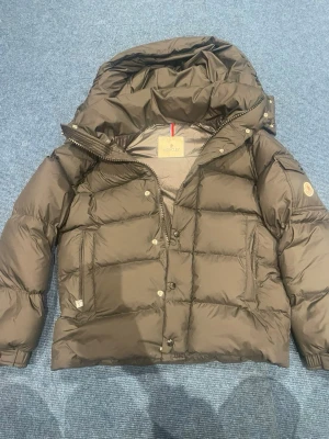 Brun dunjacka från Moncler - Snygg brun dunjacka från Moncler med stor huva och quiltad design. Jackan har dragkedja och tryckknappar framtill samt Moncler-logga på ärmen. riktigt varm tack vare dunfyllningen.