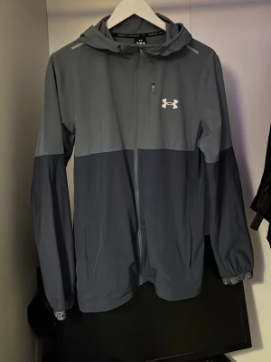 Grå vindjacka från Under Armour - Snygg tvåtonad grå vindjacka från Under Armour med huva och hel dragkedja framtill. Jackan har en vit logga på bröstet, diskreta fickor och elastiska muddar med logotypdetaljer. Tillverkad i lätt och smidigt material, perfekt för blåsiga dagar.