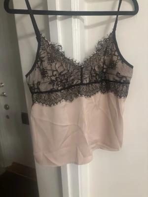 Beige linne med svart spets - Säljer ett sophiastiligt beige linne med tunna axelband och detaljerad svart spets upptill. Spetsen har blommönster och ger en lyxig känsla. Materialet är tunt och silkeslent, perfekt för att känna sig extra fin. Passar till både chill och merAttra outfits.