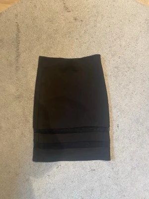 Svart kortkjol med meshdetaljer - Säljer en svart kortkjol med två genomskinliga meshpaneler längst ner. Kjolen har en tight passform och är perfekt för dig som gillar en stilren men edgy look. Materialet känns mjukt och stretchigt, vilket gör den bekväm att bära hela dagen.