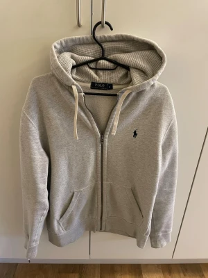 Grå hoodie från Polo Ralph Lauren - Snygg grå hoodie från Polo Ralph Lauren med dragkedja, två fickor framtill och klassisk huva med vita snören. Ikoniska Polo-loggan broderad på bröstet. Tillverkad i mjukt sweatshirtmaterial som är perfekt för chill dagar.