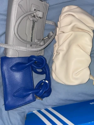 Tre trendiga handväskor i olika färger - Säljer tre snygga handväskor: en ljusgrå med detaljer och dragkedja, en krämvit med puffig, rynkad design och en klarblå med chunky handtag.