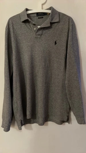 Grå långärmad piké Polo Ralph Lauren - Grå långärmad pikétröja från Polo Ralph Lauren i custom slim fit. Klassisk krage, knappar vid halsen och broderad logga på bröstet. Tillverkad i mjuk bomull för en clean och stilren look.