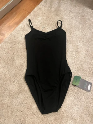 Svart body - Svart body med tunna axelband och rundad urringning. Baksidan har en djupare öppning och insidan är delvis fodrad i beige tyg. Perfekt för att skapa en clean och stilren look. Den har andväts till ballet.