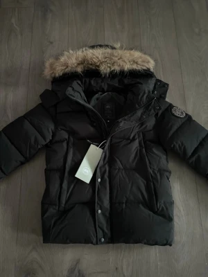 Canada Goose jacka svart - Snygg svart dunjacka från Canada Goose med avtagbar huva och fuskpäls. Jackan har en patch på ärmen, dragkedja och tryckknappar framtill samt flera fickor. Perfekt för kalla vinterdagar och riktigt varm.