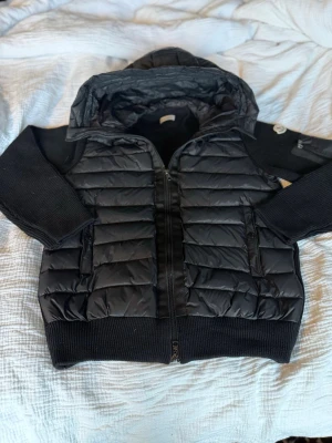 Svart pufferjacka från Moncler - Snygg svart pufferjacka från Moncler med stickade ärmar och huva. Jackan har quiltad front och rygg i glansigt material, dragkedja framtill och två fickor med dragkedja. Perfekt för våren och hösten och riktigt trendig look. Nfc Scan funkar 