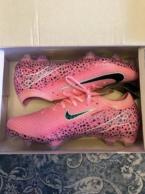 Nike rosa Mercurial fotbollsskor - Säljer ett par Nike Mercurial fotbollsskor i en riktigt snygg rosa färg med svart och vitt mönster. Skorna har coola prickdetaljer och en skinande, genomskinlig sula med texten Air Zoom. Perfekta för dig som vill sticka ut på planen och ha grymt grepp.