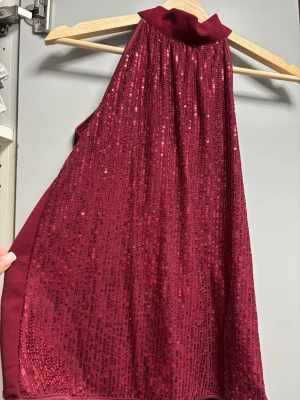 Vinröd glittrig halterneck topp - Snygg vinröd halterneck topp med glittriga paljetter framtill och knyt i nacken. Toppen är ärmlös och har en elegant, lös passform. Perfekt för dig som vill sticka ut med lite extra bling. Materialet känns som polyester och syntet.
