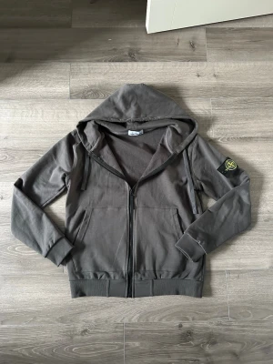 Mörkgrå hoodie från Stone Island - Säljer en mörkgrå hoodie från Stone Island med dragkedja framtill och klassisk logopatch på vänster ärm. Hoodien har två stora fickor fram, ribbade muddar och huva med dragsko. Perfekt för en avslappnad och stilren look.