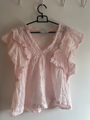 Ljusrosa volangblus med v-ringning - Supersöt ljusrosa blus med volangdetaljer och v-ringning. Blusen har korta ärmar med rynkade partier och romantisk känsla.