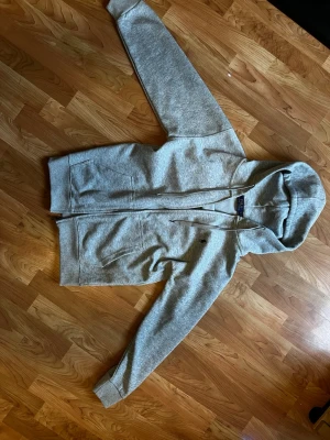 Grå hoodie från Polo Ralph Lauren - Snygg grå hoodie från Polo Ralph Lauren med dragkedja framtill och två fickor. Klassisk huva med dragsko och diskret logga på bröstet. Tillverkad i mjukt bomullsmaterial som är skönt mot huden. Perfekt för en avslappnad och stilren look. 