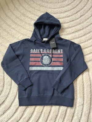 Mörkblå hoodie från Saint Laurent - Snygg mörkblå hoodie från Saint Laurent med tryck i vitt och rött framtill. Klassisk huva med dragsko och stor magficka. Tillverkad i mjukt bomullsmaterial och har ribbade muddar vid ärmslut och nederkant. Perfekt för en avslappnad och trendig look. Skickas samma dag, skriv gärna vid frågor😊