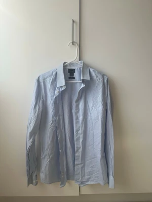 Ljusblå skjorta från H&M - Säljer en klassisk ljusblå skjorta från H&M med knappar framtill och på ärmsluten. Skjortan har en rak passform och är tillverkad i bomull, perfekt för en stilren look.