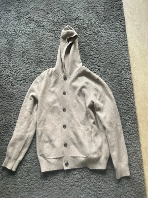 Beige hoodie med knappar - Säljer en beige hoodie med knappar framtill och två fickor. Hoodien har ribbade muddar vid ärmslut och nederkant, samt en stor huva. Materialet ser ut att vara stickat och mjukt, perfekt för en chill look.