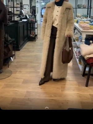 Lång beige pälsjacka - Superlång beige fuskpäls-kappa med elegant bred krage och rak, stilren passform. Ger en lyxig och exklusiv look – perfekt för dig som vill sticka ut i mängden. Kappan stängs framtill och har diskreta sidofickor. Enkel att matcha med lager-på-lager och passar både till vardag och fest. ✨ Trendig ✨ Bekväm ✨ Statement-plagg