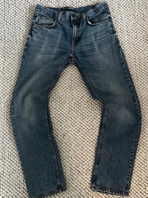 Blå raka jeans från Nudie Jeans - Säljer ett par klassiska blå jeans från Nudie Jeans med raka ben och femficksmodell. Jeansen har snygga slitningar, orange sömmar och Nudie-logga på bakfickan. Tillverkade i denim av bomull och perfekta för en avslappnad stil.
