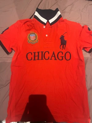 Röd Ralph Lauren Chicago t-shirt - Snygg röd t-shirt från Ralph Lauren med stor svart logga och texten CHICAGO på bröstet. Har broderad patch på vänster sida och siffran 3 på ryggen och ärmen. Klassisk pikékrage med vita och svarta detaljer. Perfekt för dig som gillar sportig stil. Gammal men perfekt skick!!