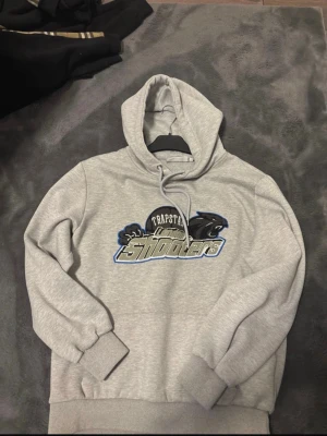 Grå Trapstar Shooters hoodie - Säljer en grå hoodie från Trapstar med stort 'Shooters'-tryck och svart pantermotiv på bröstet. Hoodien har huva med dragsko och ribbade muddar vid ärmar och nederkant. Perfekt för dig som gillar streetwear och vill sticka ut med en unik design.
