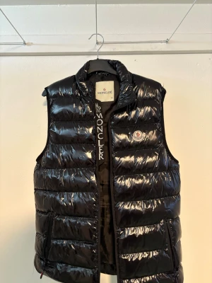 Svart dunväst från Moncler - Snygg svart dunväst från Moncler med glansig finish och hög krage. Västen har Moncler-logga på bröstet och dragkedja framtill. Perfekt för lager-på-lager och ger en sportig vibe till din outfit. Den är i gott skick förutom att det är ett litet hål vid axeln som du ser på sista bilden. Det är storlek M men den är lite liten i storleken, Kontakta vid funderingar