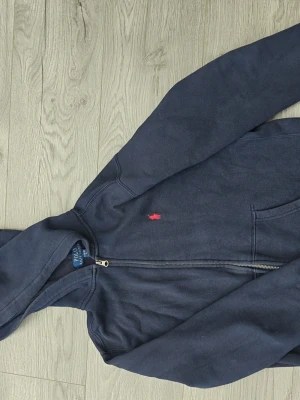 Mörkblå hoodie från Polo Ralph Lauren - Mörkblå hoodie med dragkedja från Polo Ralph Lauren. Klassisk design med huva, fickor framtill och den ikoniska röda logotypen broderad på bröstet. Denna hoodien passar för dig som är från 14 år till 16 år men storleken kan variera till skillnad från andra klädesmärken.