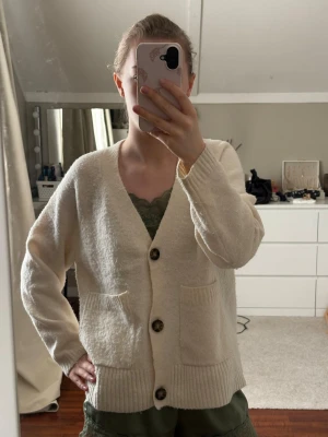 Krämvit kofta med stora knappar - Mysig krämvit kofta med v-ringning och stora bruna knappar framtill. Två rymliga fickor på framsidan och ribbade muddar vid ärmslut och nederkant. Perfekt att slänga över en topp för en avslappnad look.