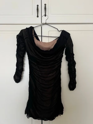 Svart meshklänning med drapering - Snygg svart kortklänning i mesh med draperade detaljer och långärmad design. Klänningen har en djup urringning med beige innerfoder som ger en cool kontrast. Perfekt för dig som vill sticka ut med en trendig look.