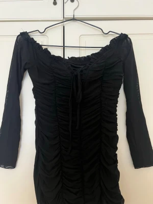 Svart offshoulder kortklänning - Trendig svart kortklänning med offshoulder-design och trekvartsärm. Klänningen har snygg rynkning framtill, snörning vid urringningen och är gjord i ett tunt, stretchigt material som ger en smickrande passform.
