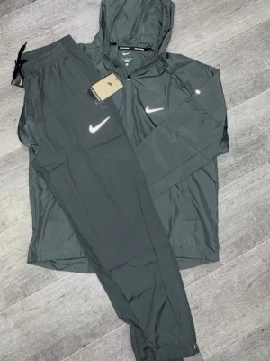 Nike Miller Tracksuit (Grå) - Snyggt svart träningsset från Nike med matchande byxor och jacka med huva. Setet har vita Nike-loggor på både jackan och byxorna. Jackan har dragkedja framtill och byxorna har resår i midjan. Perfekt för träning eller chill.