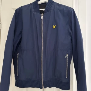 Marinblå bomberjacka från Lyle & Scott - Snygg marinblå bomberjacka från Lyle & Scott med gul logga på bröstet. Jackan har silverfärgade dragkedjor, ribbade muddar vid ärmslut och nederkant samt två fickor framtill. Klassisk bombermodell i lätt material, perfekt för en stilren look.