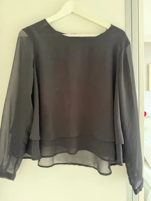 Svart transparent blus från Ally Bell - Snygg svart blus från Ally Bell i transparent, lätt material. Blusen har långa ärmar och ett lager-på-lager-utseende med rund halsringning. Perfekt att styla med jeans eller kjol för en trendig look.
