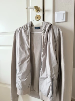 Ljusgrå vindjacka med huva från Herno - Snygg ljusgrå vindjacka från Herno med huva och dragkedja framtill. Jackan har stickade ärmar och muddar samt två fickor med dragkedja. Perfekt för dig som vill ha en stilren och sportig look. Nypris ca 6000