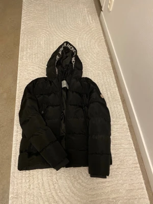 Svart dunjacka från Moncler med huva - Säljer en svart dunjacka från Moncler med huva och dragkedja. Jackan har Moncler-logga på ärmen och textdetaljer på huvan. Puffig modell som håller dig varm under kalla dagar. Perfekt för dig som vill ha både stil och funktion.